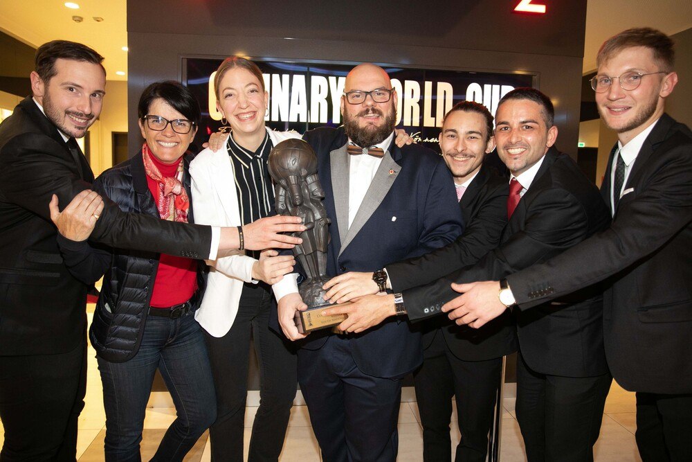 Teamchef Philipp Stohner (Vizepräsident VKÖ), Petra Nocker Schwarzenbacher (Bundesspartenobfrau in der Wirtschaftskammer Österreich), Agnes Karrasch, VKÖ-Präsident Mike P.Pansi, Celino Waldner, Trainer Alexander Forbes (Vizepräsident VKÖ) und Thomas Penz (v.l.) präsentieren die 25 Kilogramm schwere Bronze-Trophäe – den „Heiligen Gral der Köche“.
© VKÖ Teamchef Philipp Stohner (Vizepräsident VKÖ), Petra Nocker Schwarzenbacher (Bundesspartenobfrau in der Wirtschaftskammer Österreich), Agnes Karrasch, VKÖ-Präsident Mike P.Pansi, Celino Waldner, Trainer Alexander Forbes (Vizepräsident VKÖ) und Thomas Penz (v.l.) präsentieren die 25 Kilogramm schwere Bronze-Trophäe – den „Heiligen Gral der Köche“.