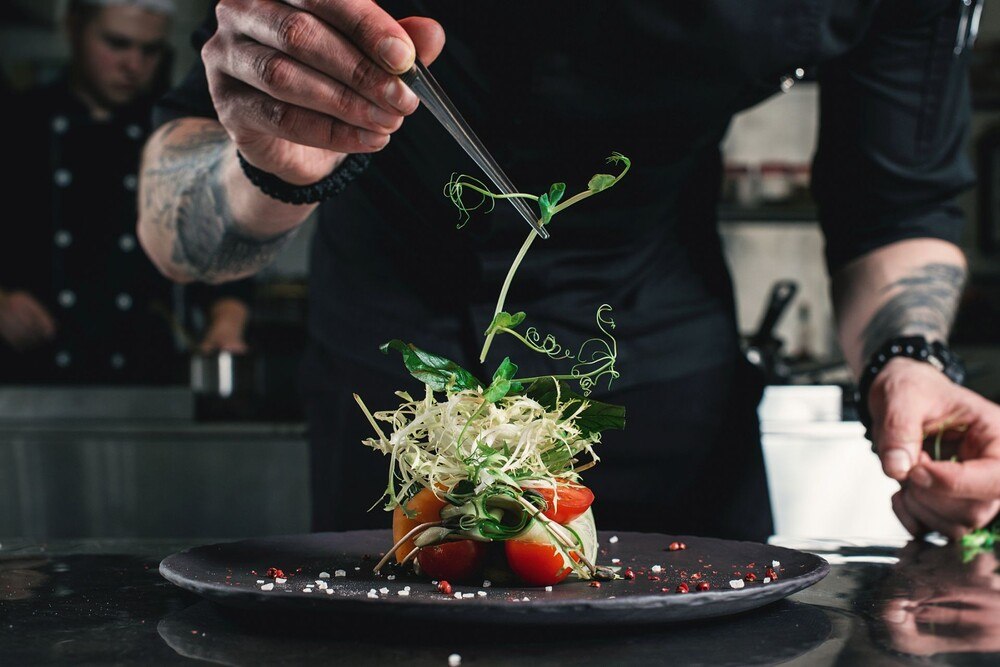 © Getty Images / MykolaSenyuk
Ein Küchenchef mit tätowierten Unterarmen richtet Salat auf einem schwarzen Teller mit der Pinzette an. Ein Küchenchef mit tätowierten Unterarmen richtet Salat auf einem schwarzen Teller mit der Pinzette an.