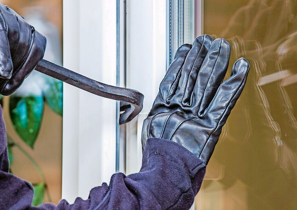 Ein Einbrecher mit Lederhandschuhen, bricht ein gekipptes Fenster mithilfe eines Brecheisens auf