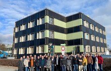 Eine Gruppe an Menschen steht vor einem grün-schwarzen Gebäude.