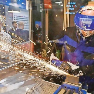 © Koelnmesse/Eisenwarenmesse Ein mit Schutzkleidung ausgerüsteter Mann flext durch ein Stück Metall sodass die Funken sprühen. Im Hintergrund betrachtet ihn jemand aufmerksam durch eine Glasscheibe