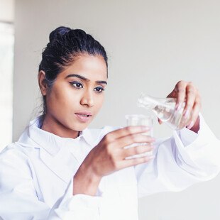 © LiudmylaSupynska - iStock / Getty Images Plus Junge indische Studentin der Biotechnologie gießt Wasser in einen Kolben für ein wissenschaftliches Experiment