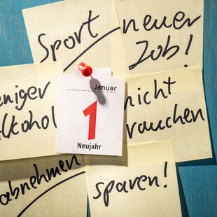 © iStock / Getty Images Plus / gopixa Das Kalenderblatt des ersten Januars ist auf eine blaue Wand aufgepinnt. Rundherum hängen Post-its mit Neujahrsvorsätzen.