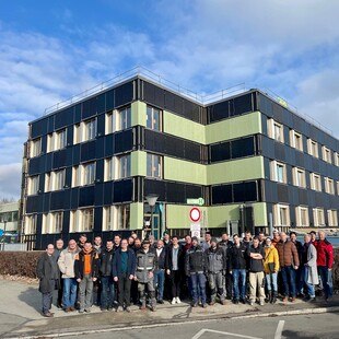 Eine Gruppe an Menschen steht vor einem grün-schwarzen Gebäude.